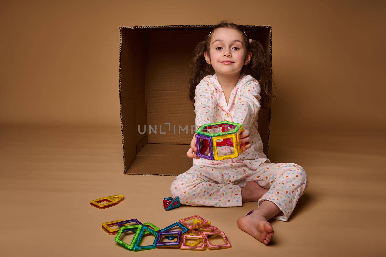 Adorable Caucasian child girl holding in her outstretched hands a built model from colorful magnetic constructor blocks and looking at camera isolated over beige background with copy ad space