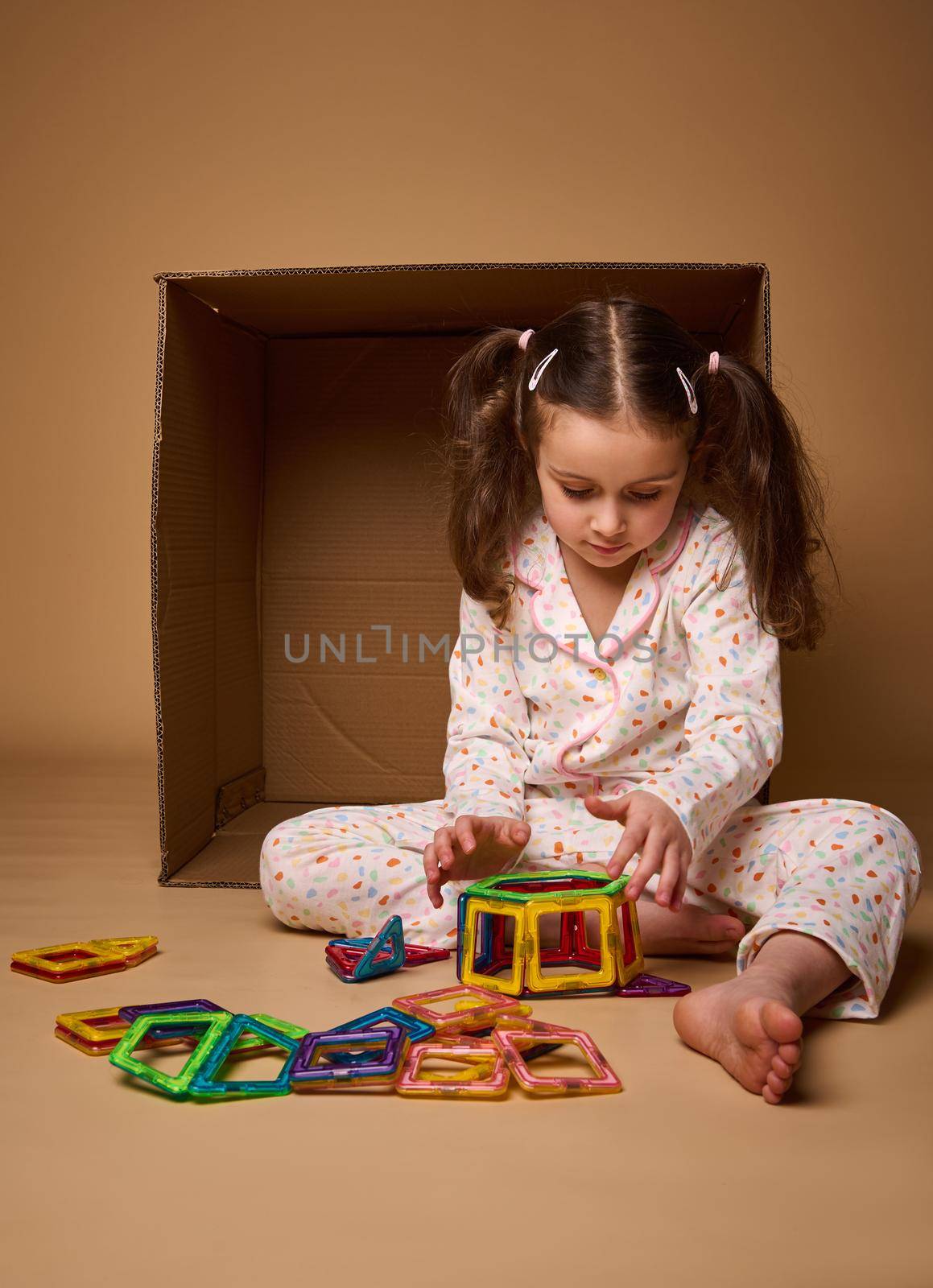 European little girl with two ponytails in pajamas playing with colorful magnetic constructor toy on beige background with copy ad space