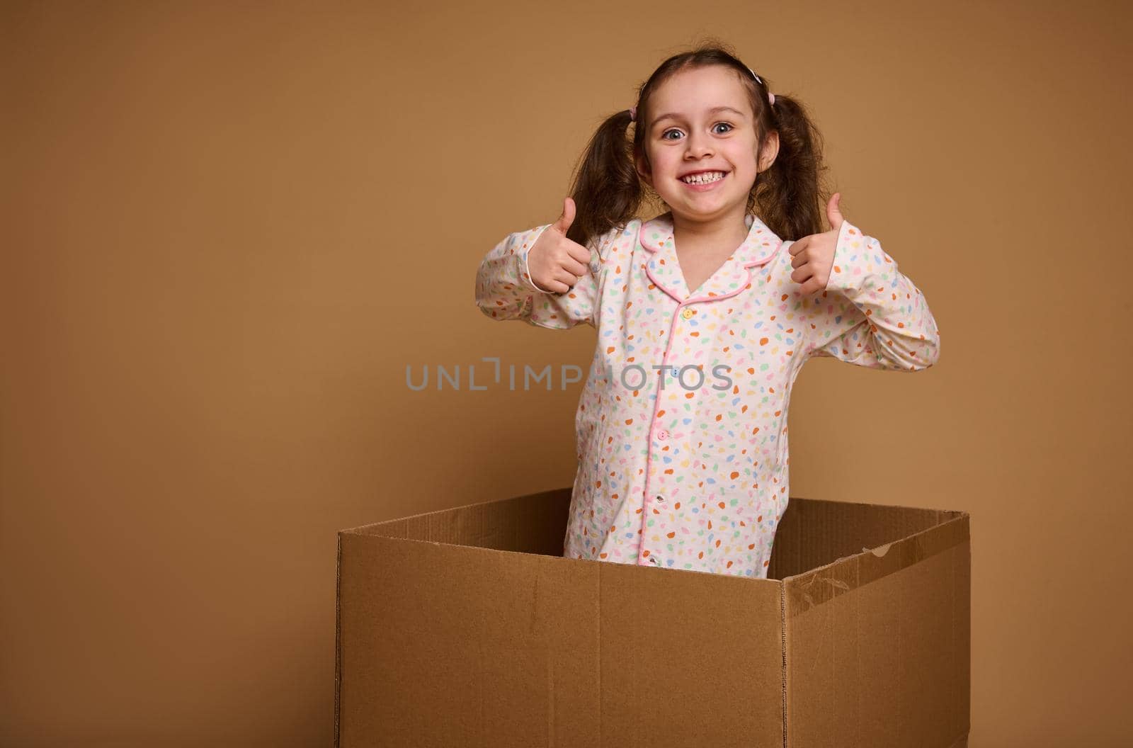 Charming cheerful kid, pretty Caucasian baby girl with two ponytails showing thumbs up, smiles looking at camera being inside a cardboard box against a beige background with copy ad space by artgf