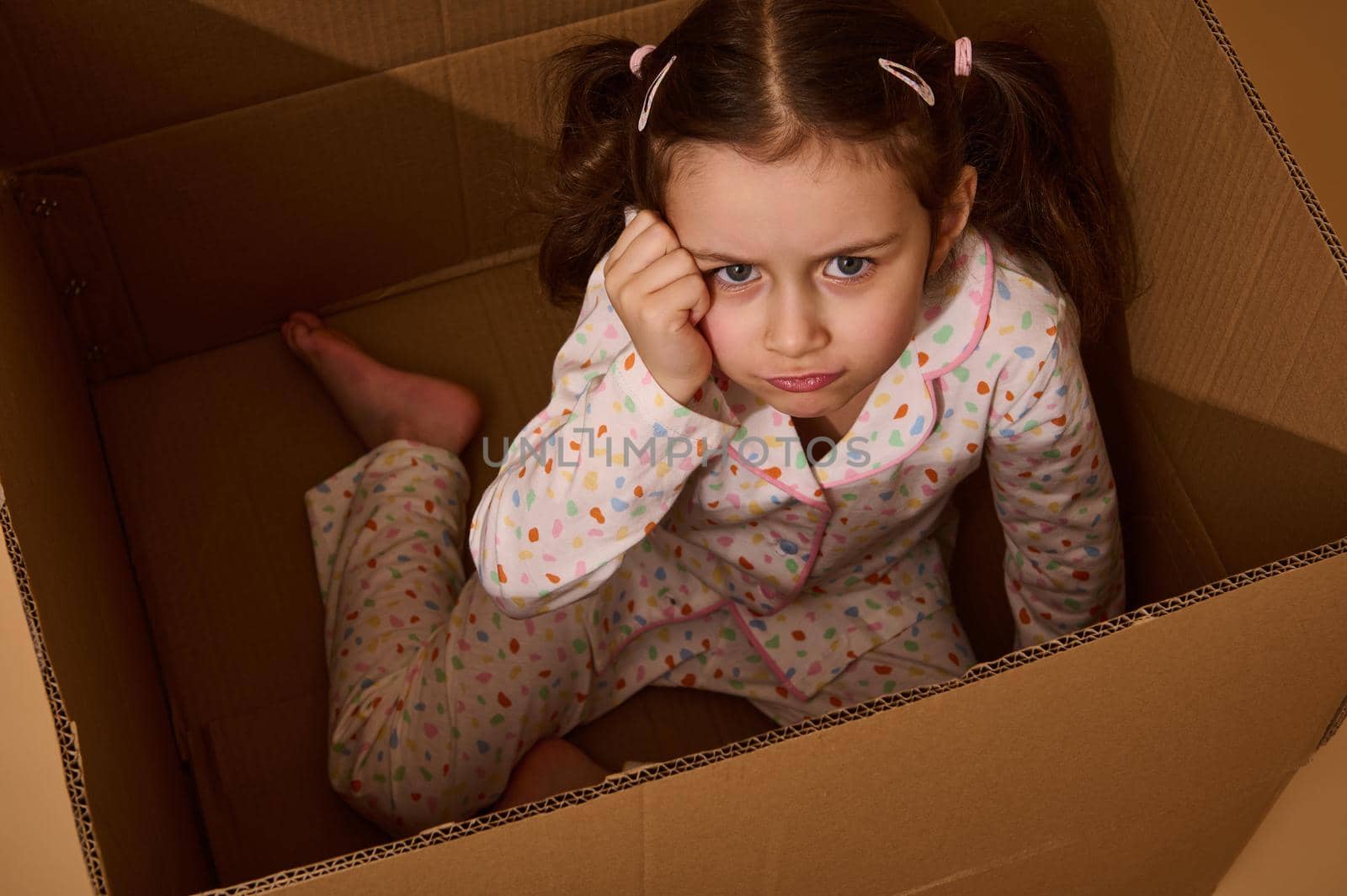 Studio shot of a beautiful upset little child girl looking at the camera in disappointment while inside a cardboard box. Emotional studio portrait against beige background with copy ad space
