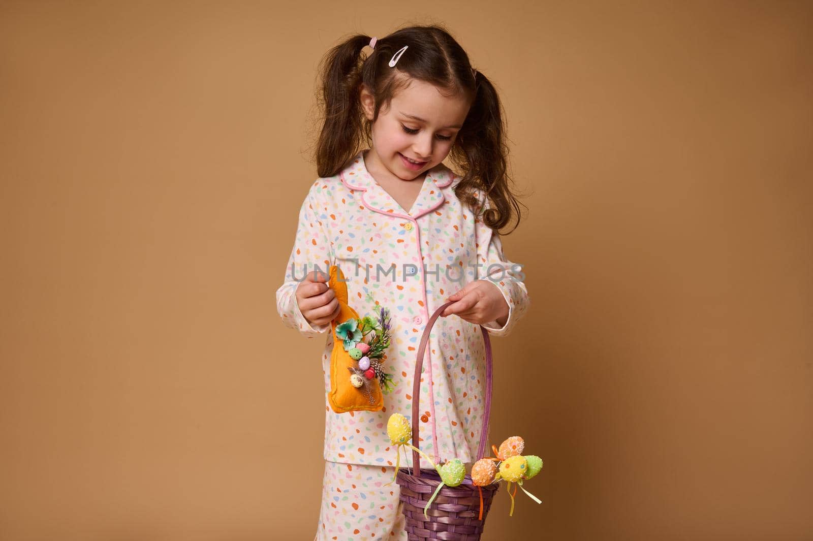 Cheerful adorable Caucasian baby girl in pajamas holding a rabbit toy and colorful decoration of Easter eggs, smiles posing to camera, isolated over beige background with copy ad space by artgf