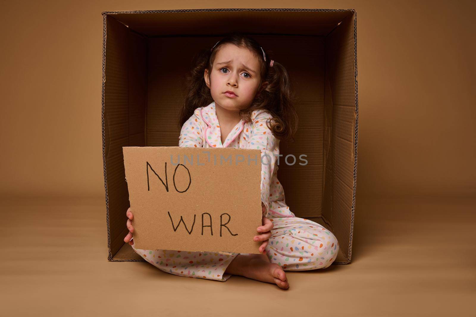 Upset European little girl sitting inside a cardboard box and showing a Stop the War poster. The concept of a ceasefire, problems of migrants and refugees during the military conflict by artgf