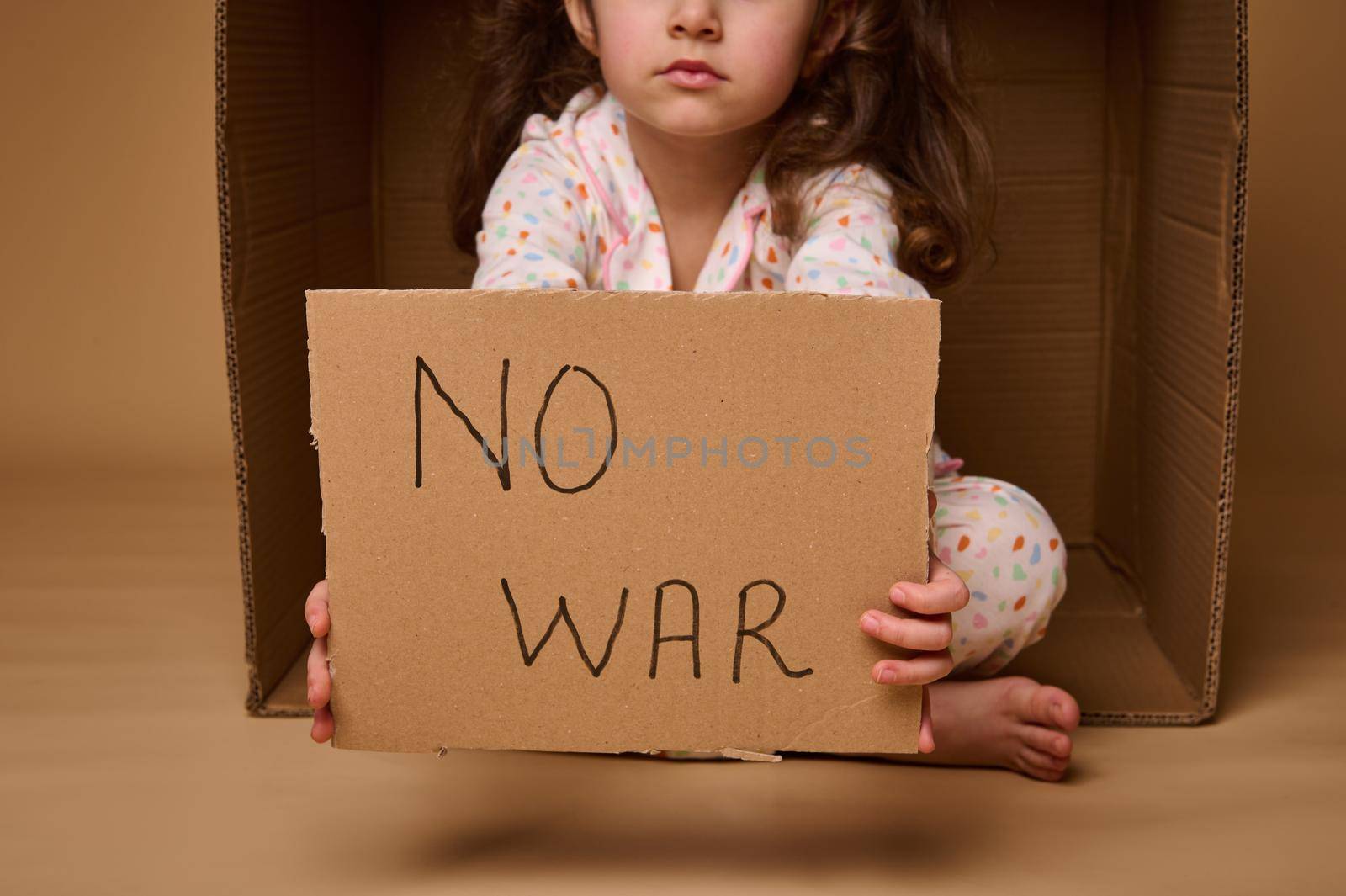 No War written in a cardboard poster in the hands of European child little girl refugee or migrant during political and military conflicts in the country, isolated over beige background, copy ad space