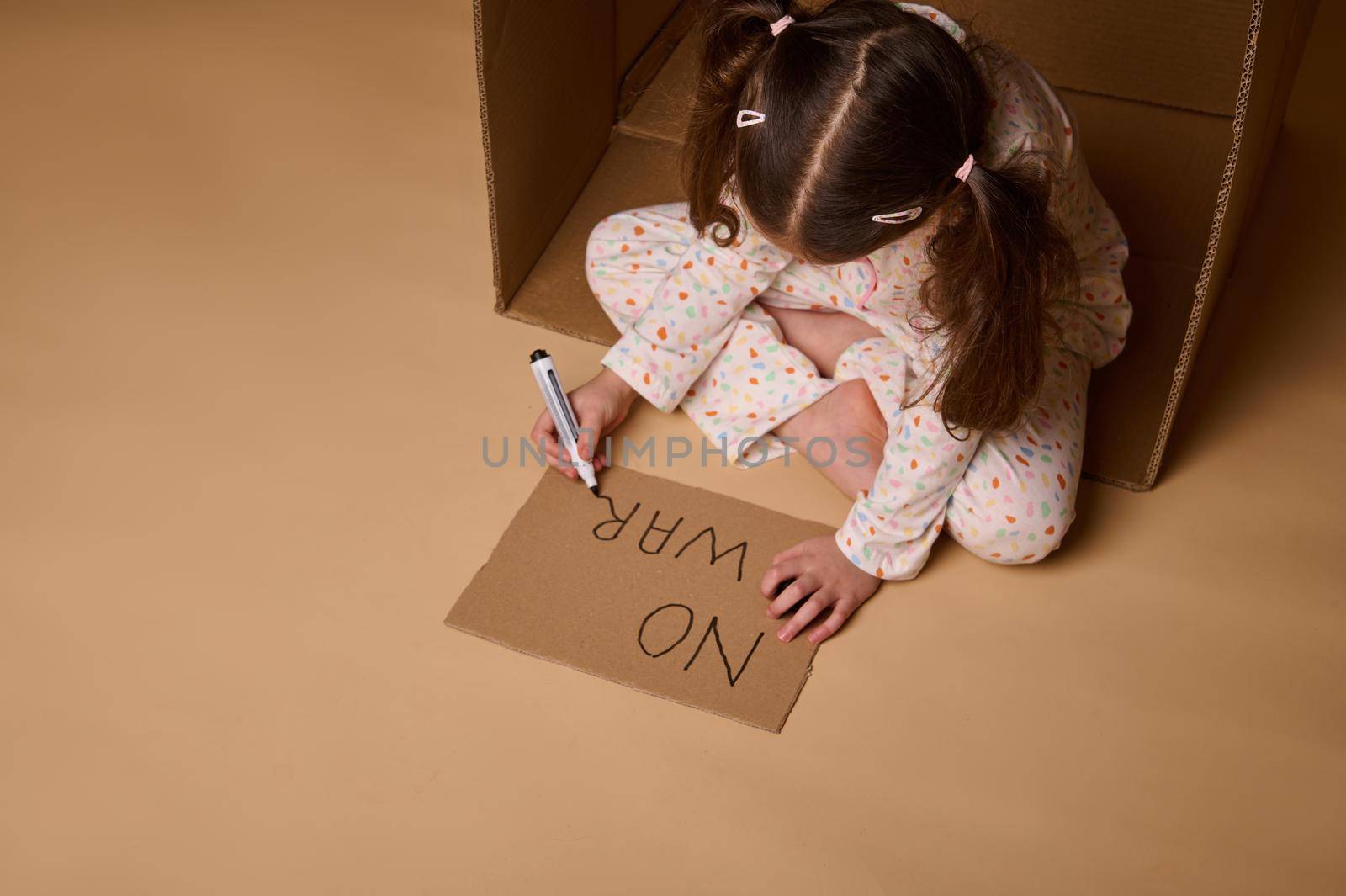 Top view of child in pajamas writing No War on a cardboard poster inside a box, hiding from the military and political conflict. Concept of refugees and immigrants losing their home during war by artgf