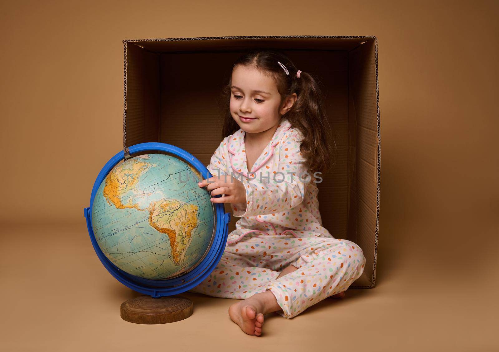 Adorable baby girl in pajamas sitting ahead a cardboard box and examining the Earth globe, isolated over beige background with copy space. Love and save planet, environment conservation concept by artgf
