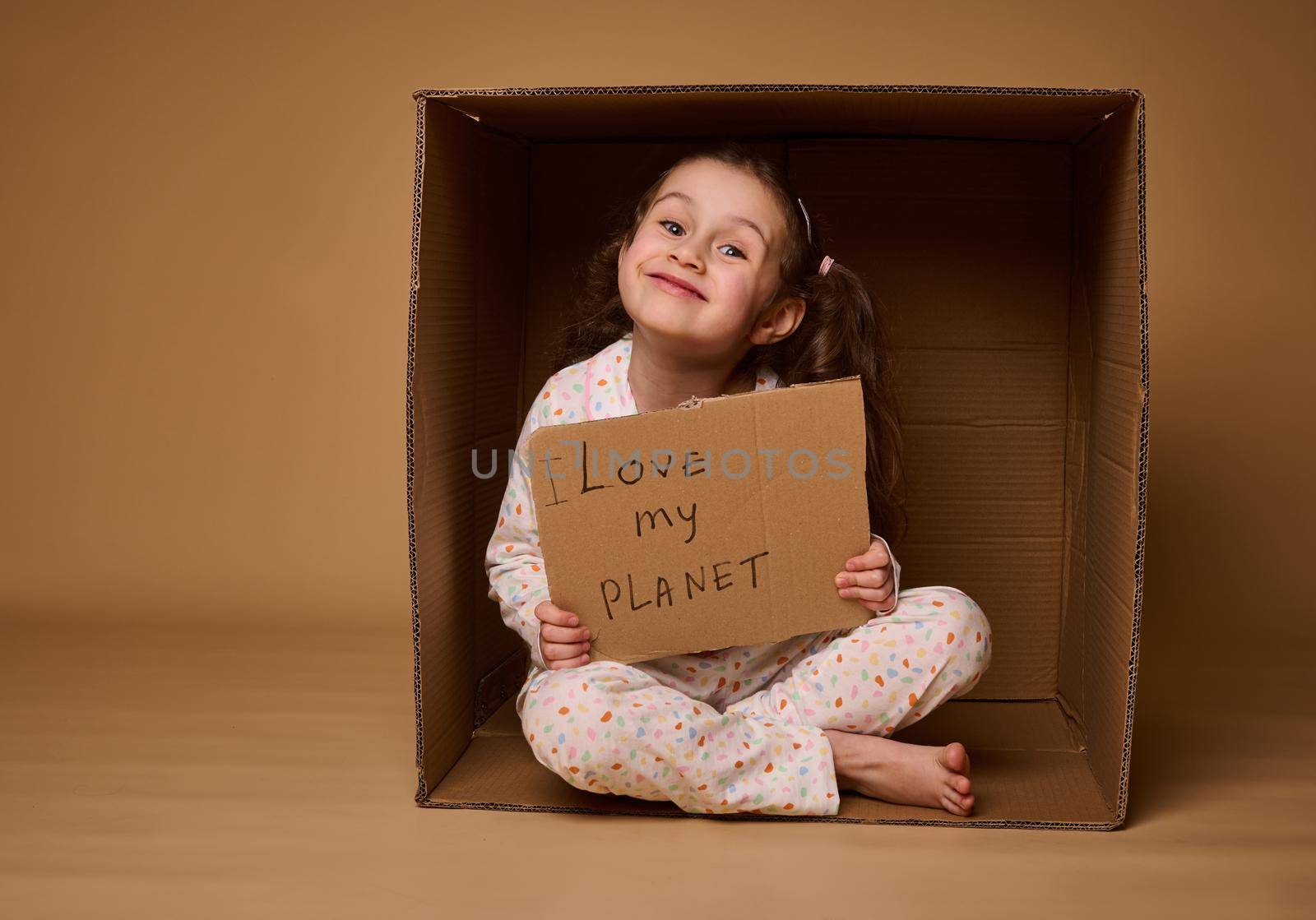 Adorable little European girl activist holding a poster with Love my Planet, sitting inside a cardboard box in lotus pose and smiling cutely looking at camera, against beige background with copy space