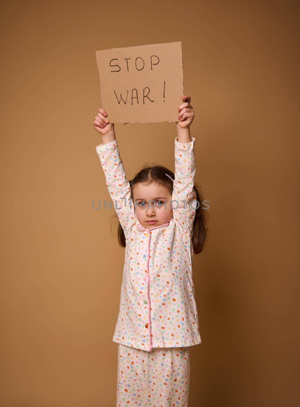 Studio shot of an adorable upset 4 years old kid, sad little European girl in pajamas holding Stop War social message cardboard poster standing on beige background with ad copy space