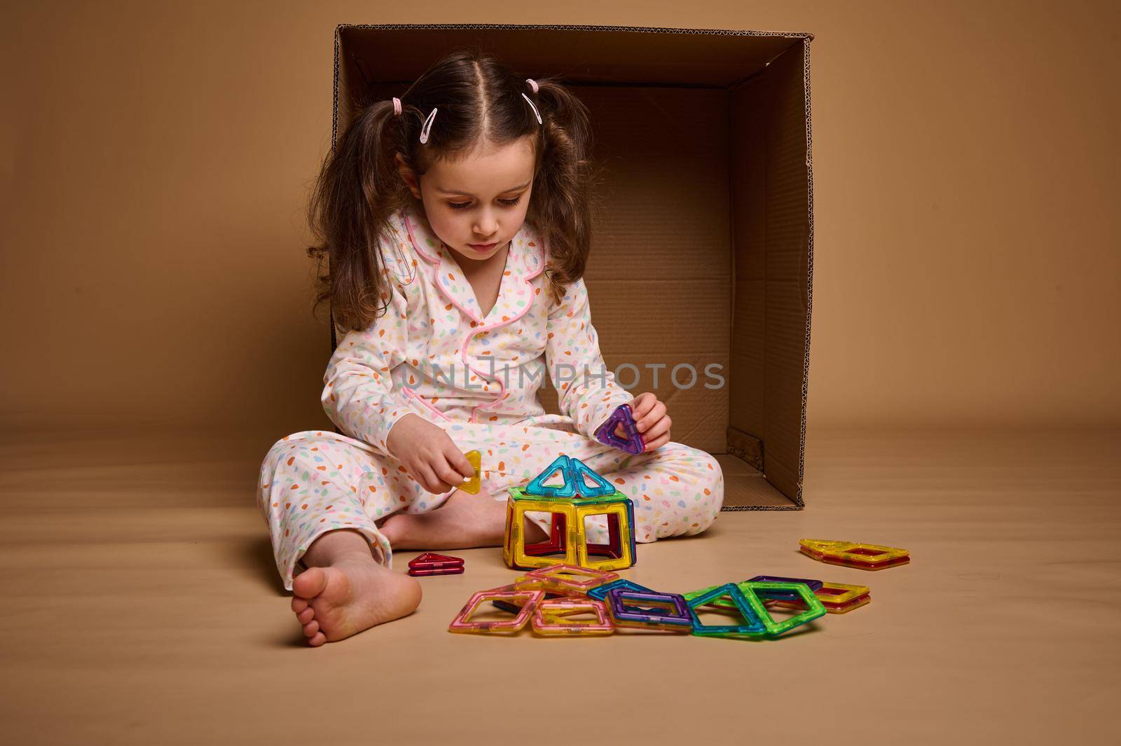 Caucasian baby girl wearing a pajamas, builds with magnetic constructor, sitting ahead a cardboard box, isolated over beige background with copy space for advertisement