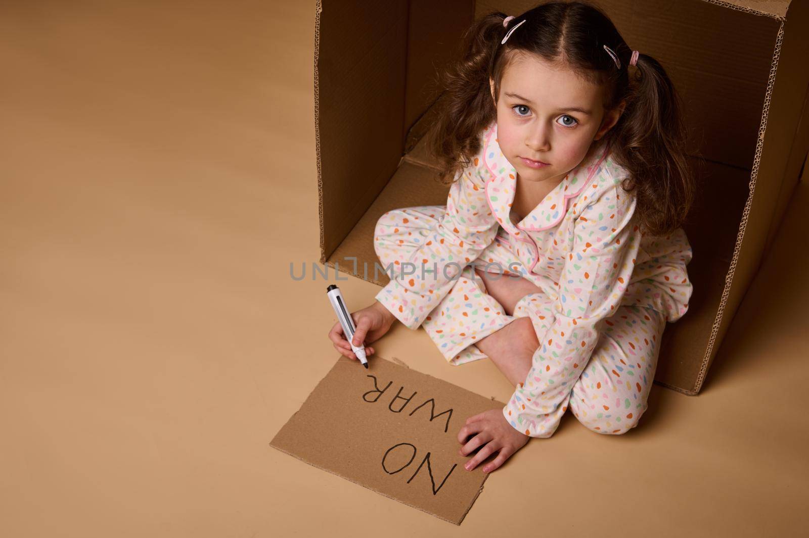 Top view little girl in pajamas writing No War on poster while sitting inside cardboard box, hiding from military and political conflict. Concept of refugees, immigrants losing their home during war by artgf