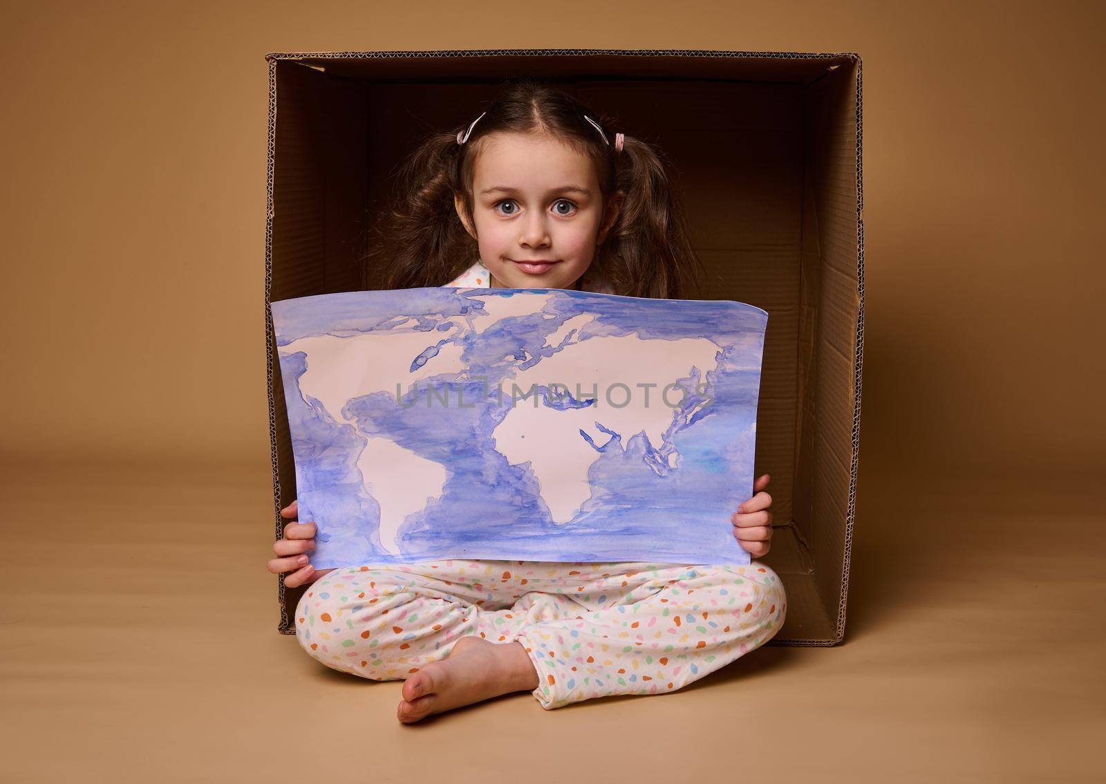 Beautiful little girl with two ponytails sitting inside a cardboard box with a poster- a painted image with World map, isolated over beige background.Environment conservation and save planet concept by artgf