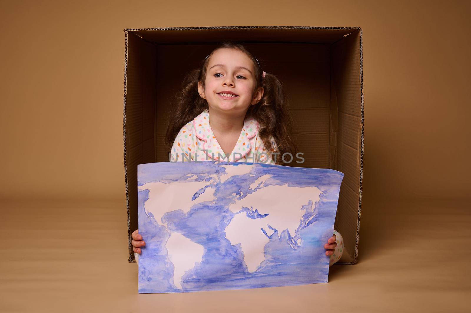 Beautiful little girl with two ponytails sitting inside a cardboard box with a poster- a painted image with World map, isolated over beige background.Environment conservation and save planet concept