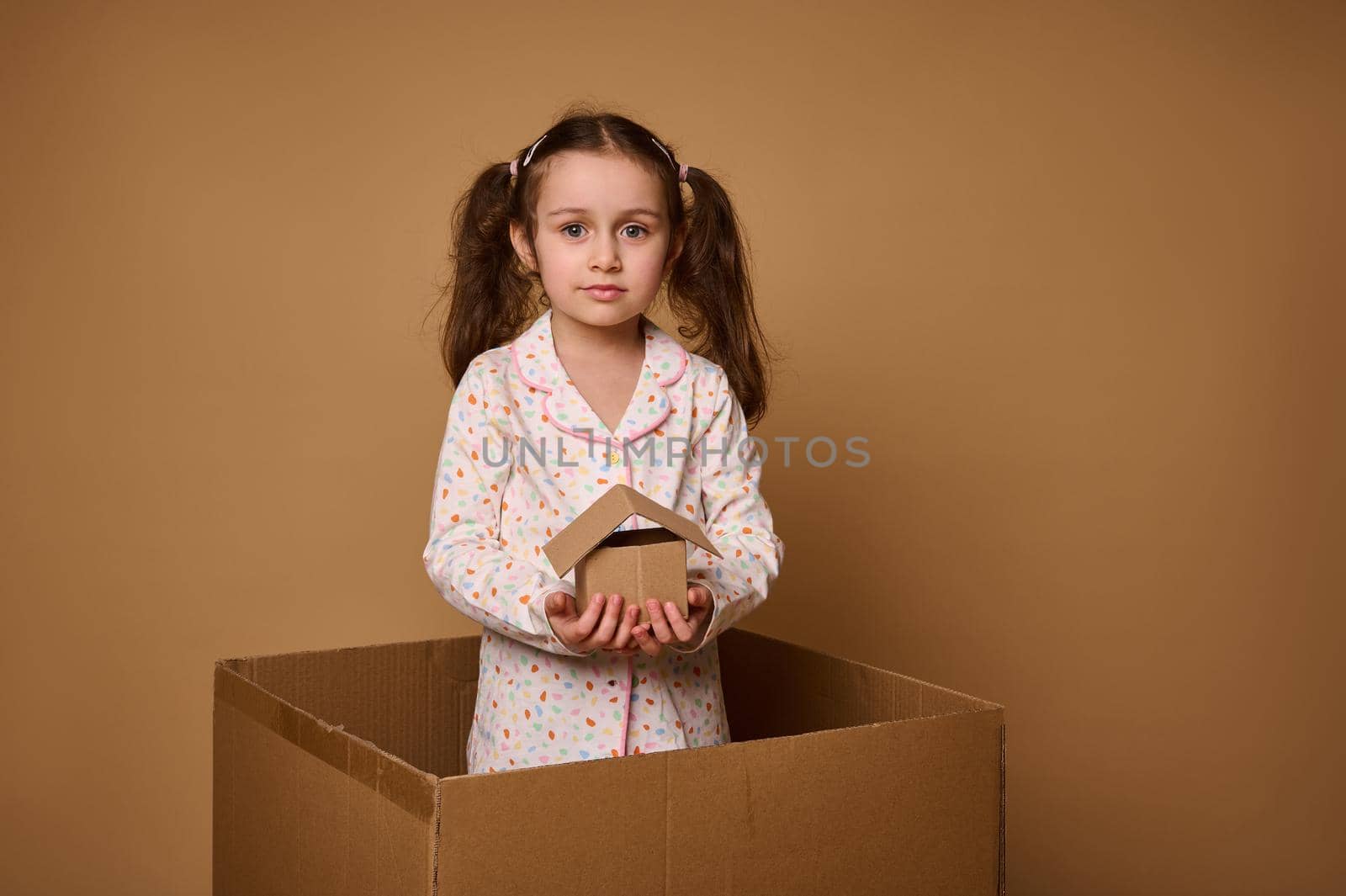 Little Caucasian girl, adorable child in pajamas holds a craft cardboard house model being inside a box, against beige background with copy space for ads. The concept of investment, housing