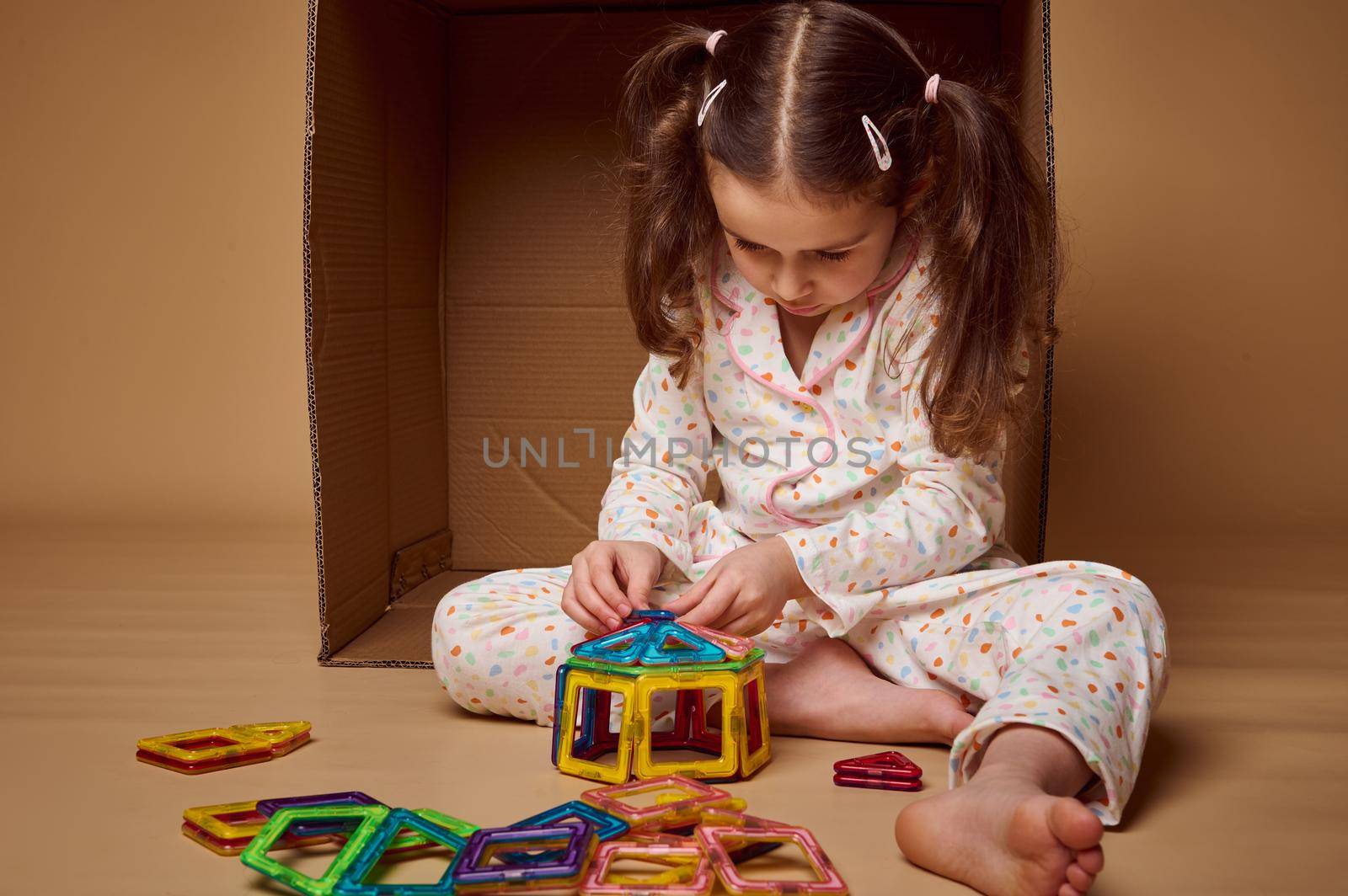 Concentrated little preschool girl, Caucasian child in pajamas sitting ahead a cardboard box and playing with magnetic constructor. Kids entertainment and fine motor kills development