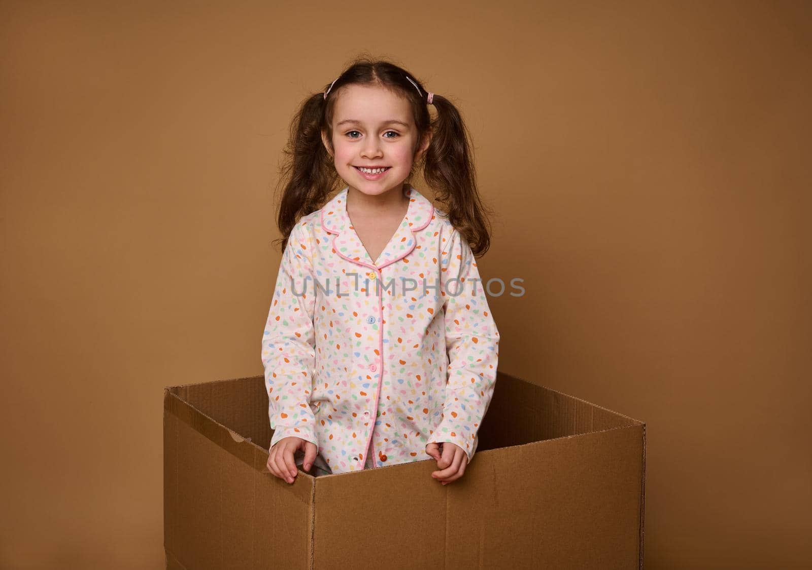 Adorable child, European 4 years old baby girl with two wavy ponytails in a bright pajamas looking at camera standing inside a cardboard box, isolated over beige background with copy space for ads