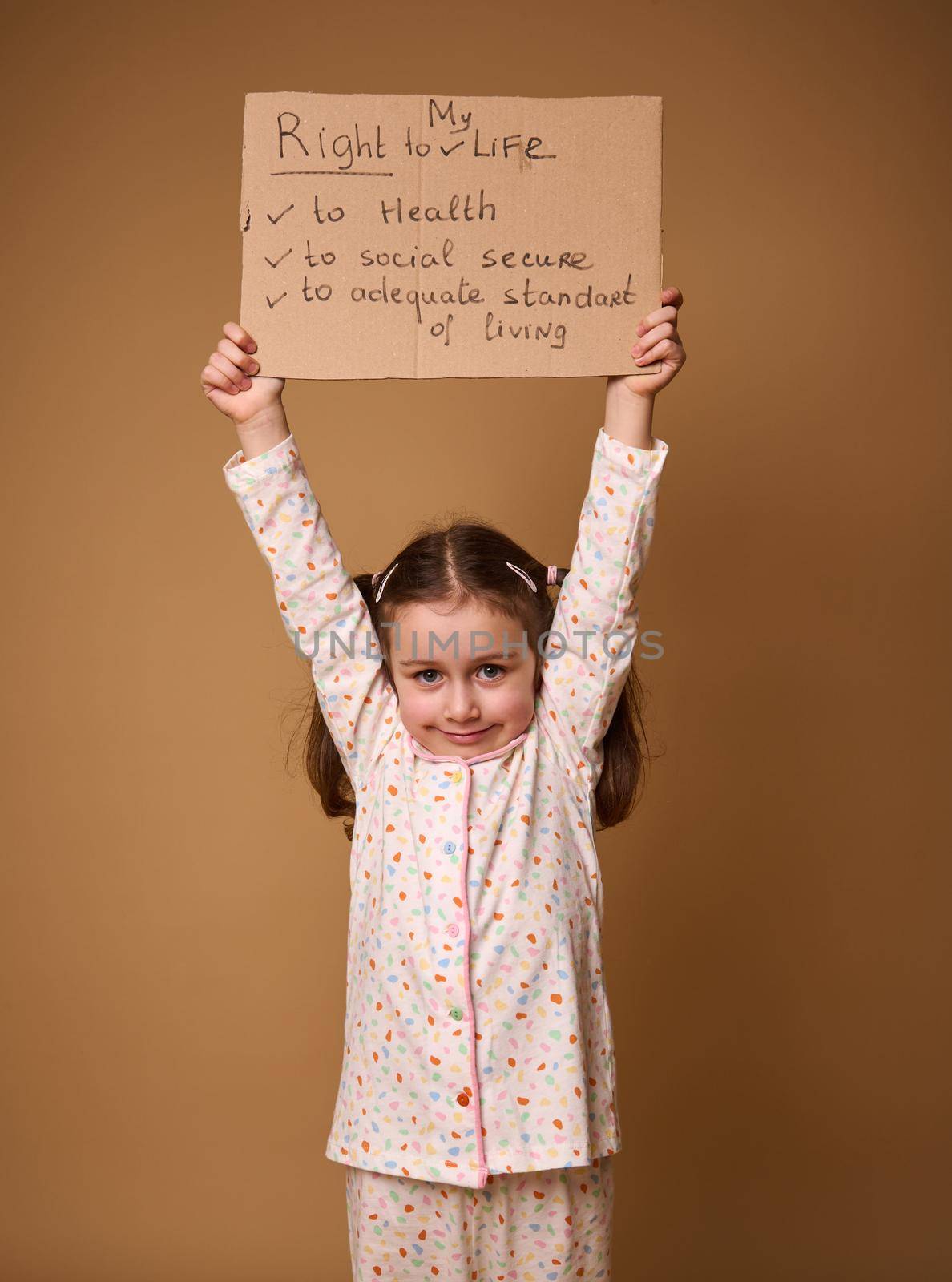 Adorable Caucasian preschool girl holding cardboard poster promoting children's rights to an adequate standard of living, social secure and health care, isolated on a beige background with copy space by artgf