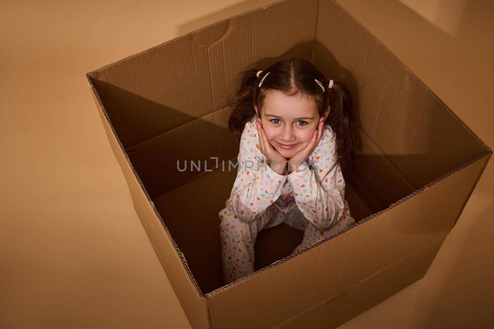 Top view of a charming little Caucasian girl sitting inside a cardboard box and looking at camera, isolated over beige background with copy space by artgf