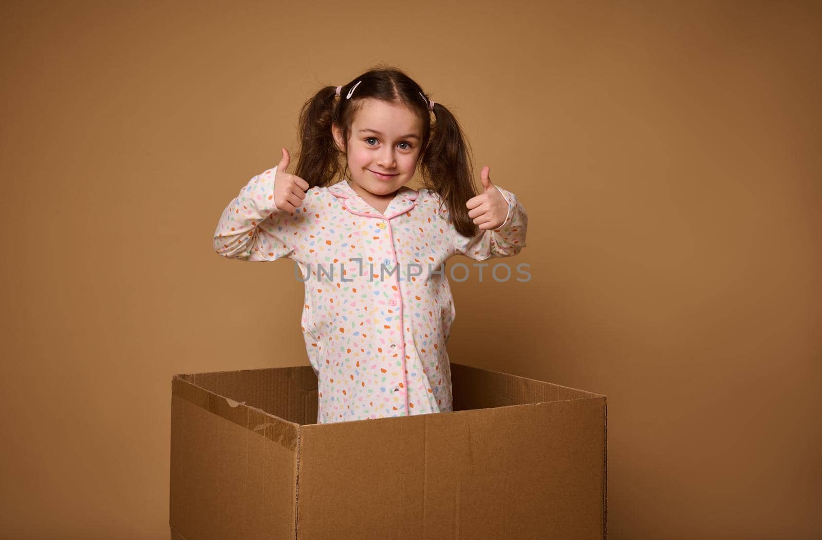 Charming cheerful child, pretty baby girl showing thumbs up, being inside a cardboard box against a beige background with copy ad space by artgf