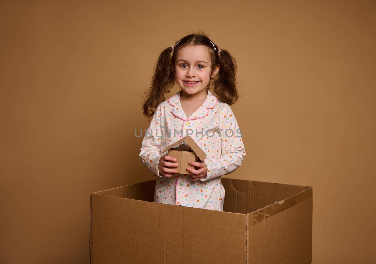 Little Caucasian girl, adorable child in pajamas holds a craft cardboard house model being inside a box, against beige background with copy space for ads. The concept of investment, housing by artgf