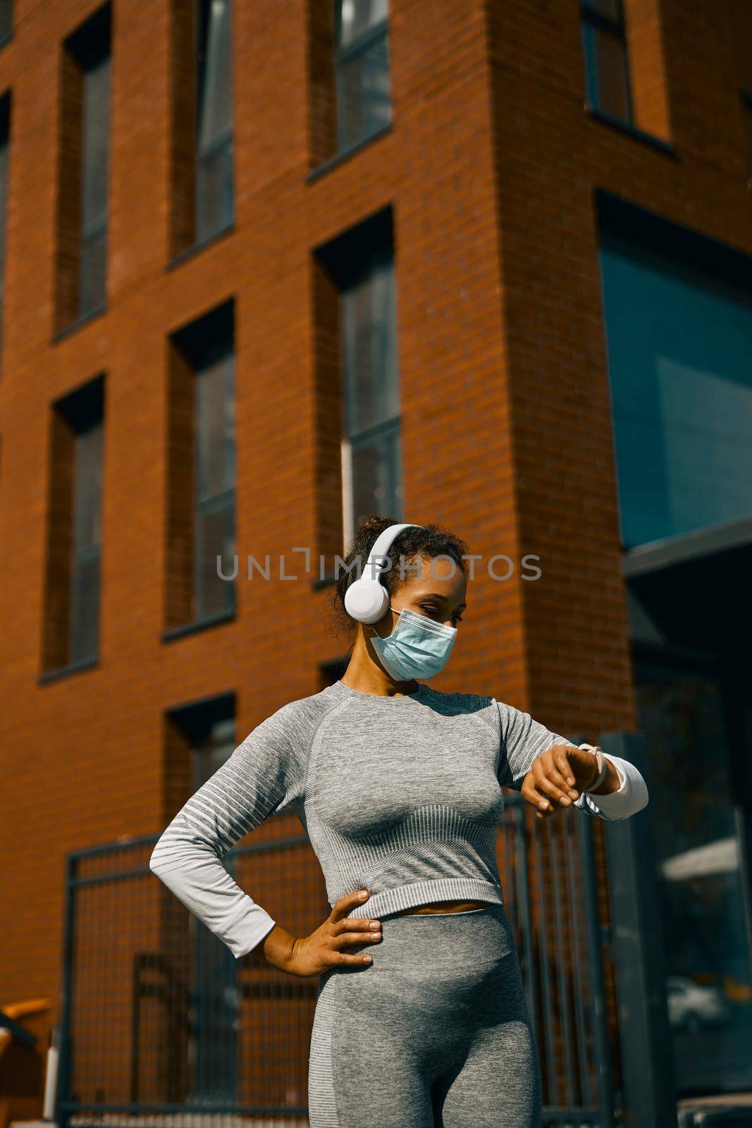 Woman in light gray sportswear and medical mask having a break during training on the street