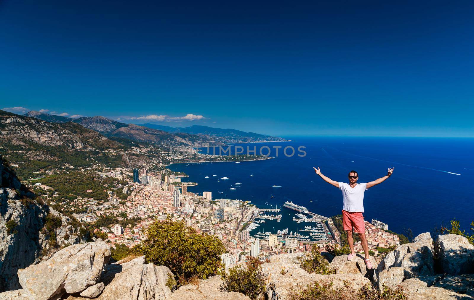 A handsome young man stands and jubilantly with his hands raised on a mountain with the name Of a Head of dog, the Principality of Monaco in the background in clear sunny weather. High quality photo