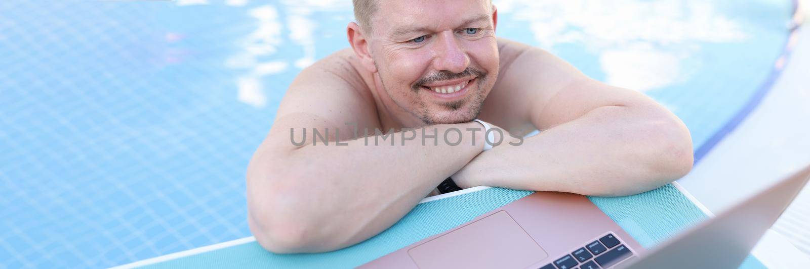 Man lying on side of swimming pool and looking at laptop screen by kuprevich