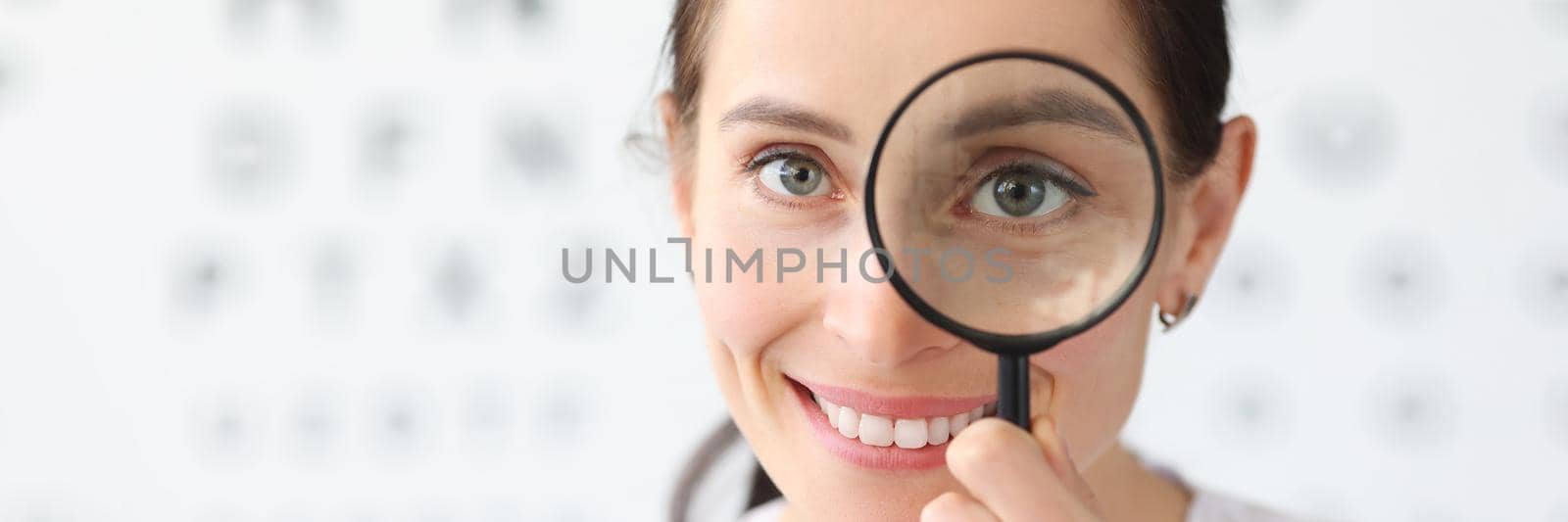 Woman optometrist holding magnifying glass in eyes against background of vision examination table. Correction of myopia concept