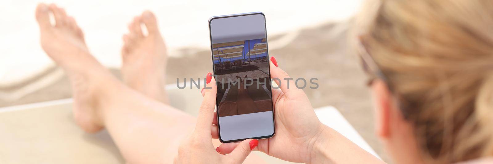 Young woman lying on sun lounger on beach and taking pictures of her feet on mobile phone closeup. Photo on social networks concept