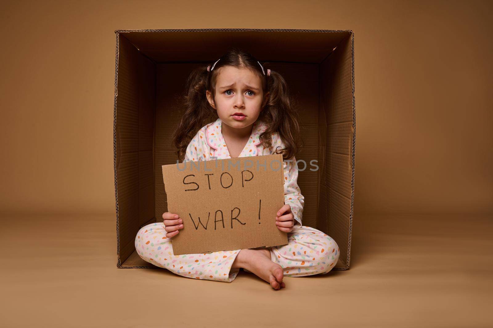 Upset European little girl sitting inside a cardboard box and showing a Stop the War poster. The concept of a ceasefire, problems of migrants and refugees during the military conflict