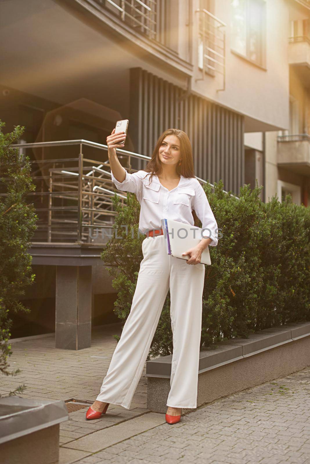 Beautiful young caucasian businesswoman in a white clothes with a laptop and notebook outdoor near business centre.