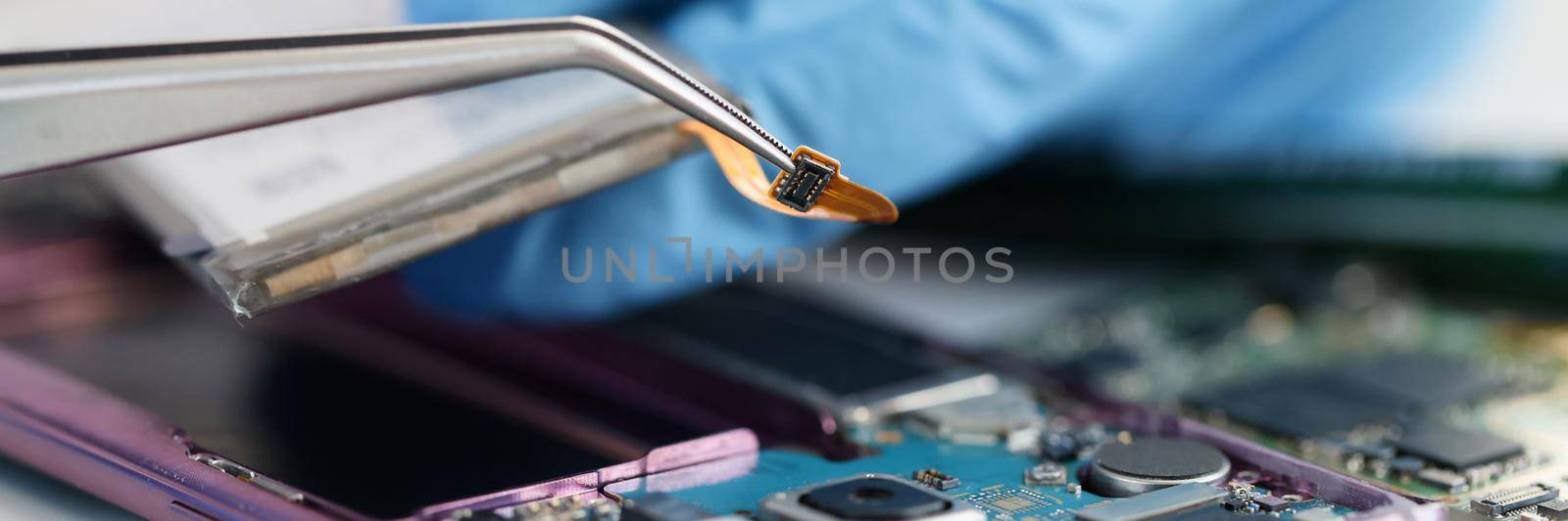 Master in rubber gloves holding microchip over broken phone in workshop. Mobile phone repair concept