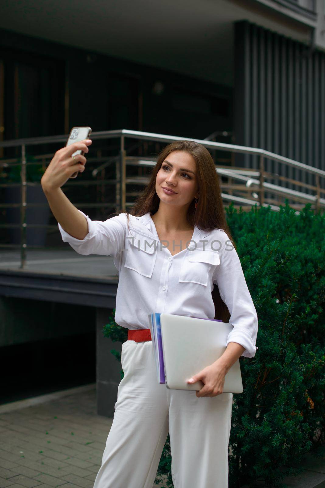 Beautiful young caucasian businesswoman in a white clothes with a laptop and notebook outdoor near business centre.