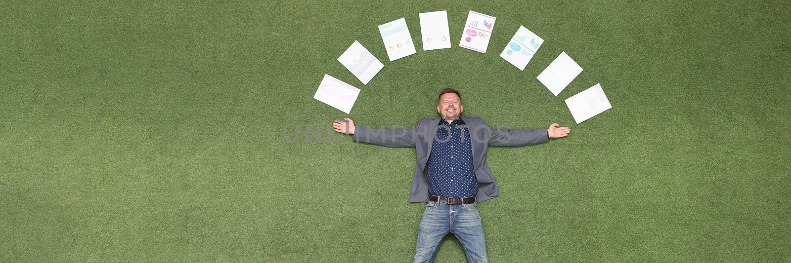 Young businessman lying on green grass with documents around his head top view. Successful business deals concept