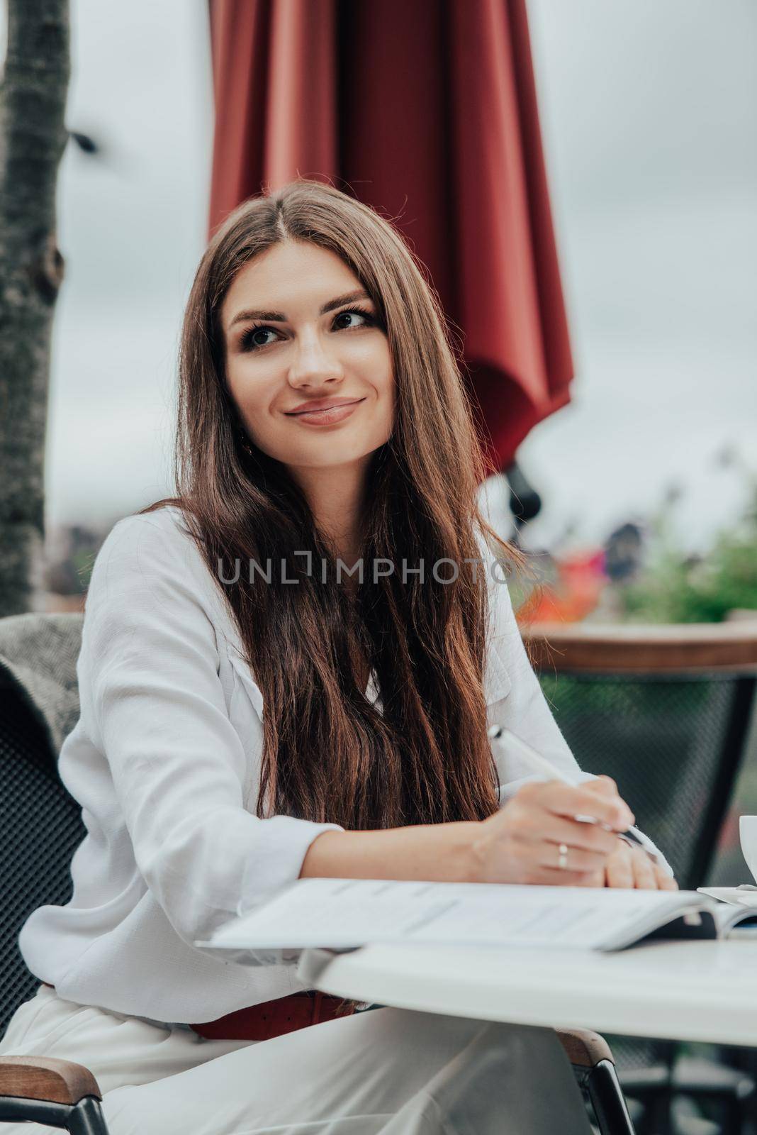 Smiling young dark-haired businesswoman makes notes in a notebook in a cafe on a summer day. Business, e-learning, freelance concept. laptop on a table.