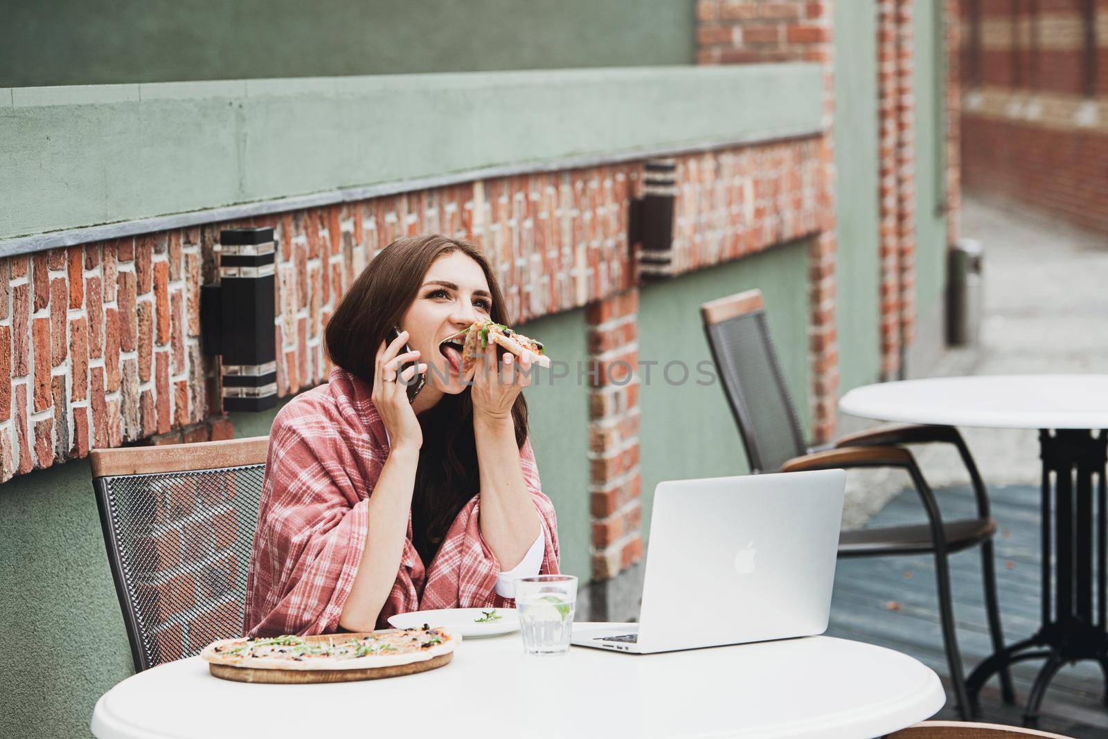 Young freelancer communicates using smartphone while eating pizza in cafe by Ashtray25