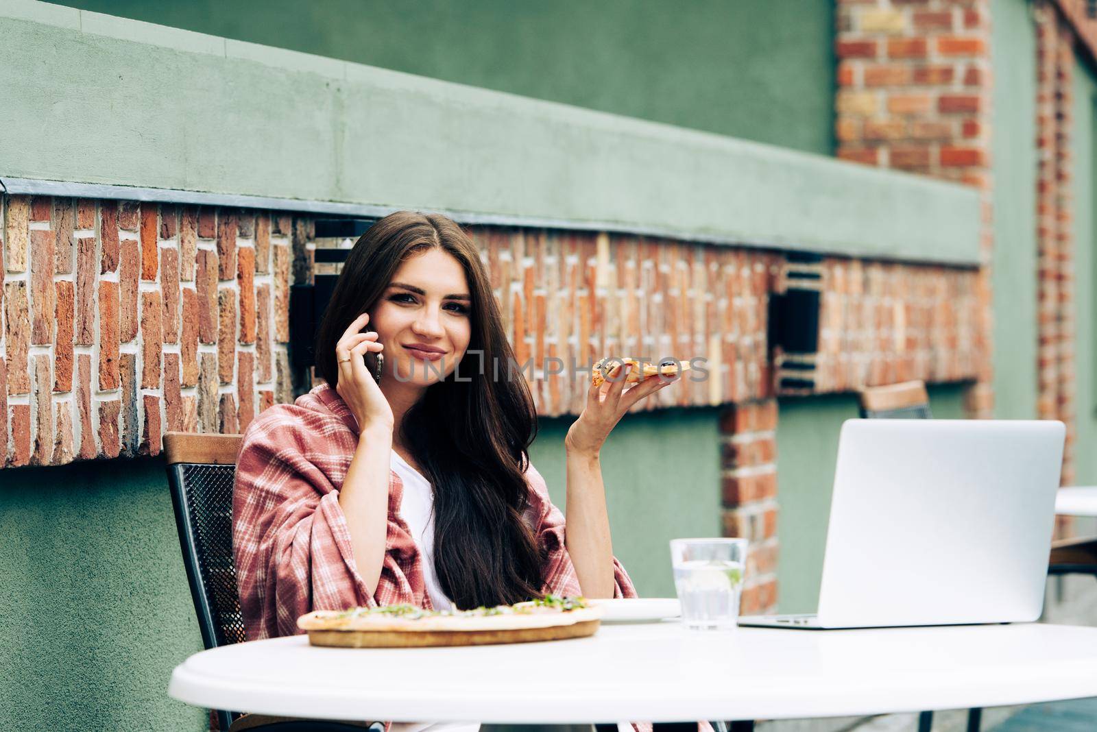 Young freelancer communicates using smartphone while eating pizza in cafe by Ashtray25