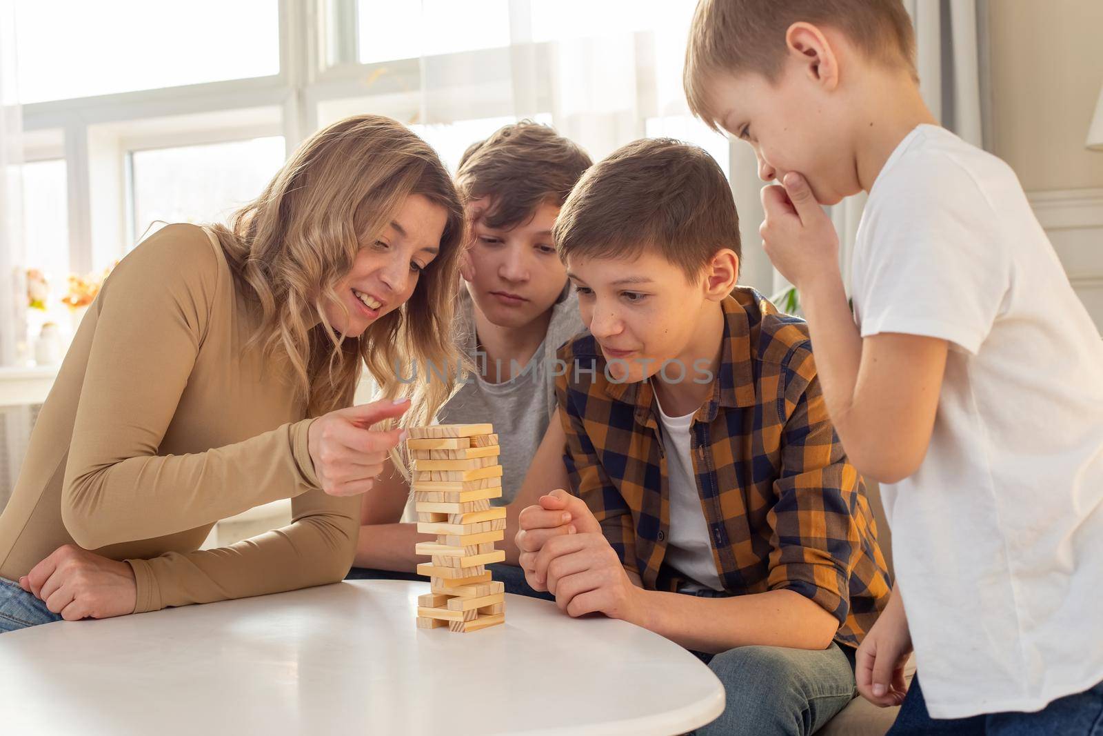 Three boys and woman, in a room, playing a board game made of wooden rectangular blocks, pulling pieces out of the tower. Top view