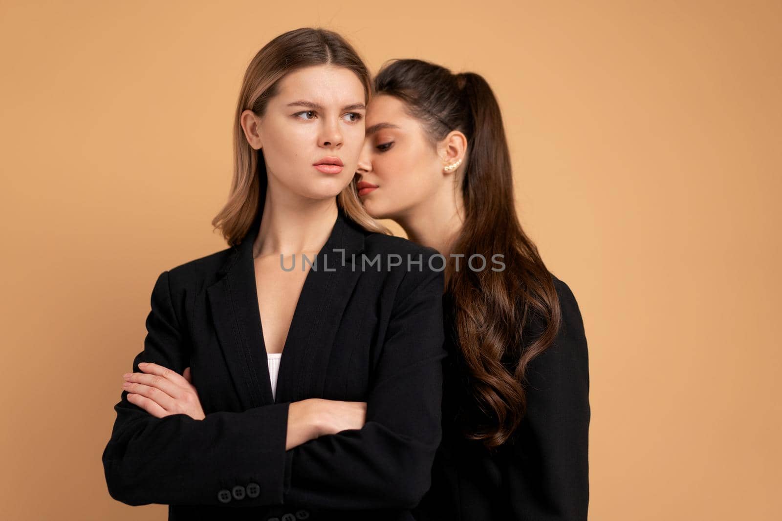 Two young business woman dressed black suit standing studio orange color background. One woman looking looking away arms crossed on chest other woman put her head on her shoulder. Friends support