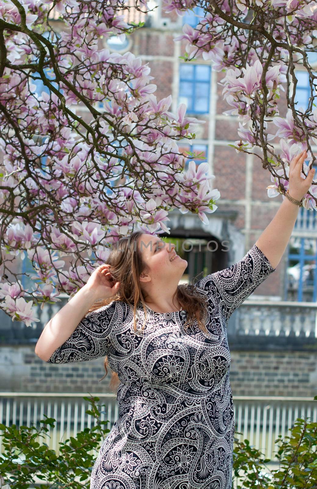 A plump, confident woman stands under a blooming magnolia and smiles. young millennial woman with brown curly hair smiling and waiting for a friend for a date. A full girl enjoys flowering and spring.