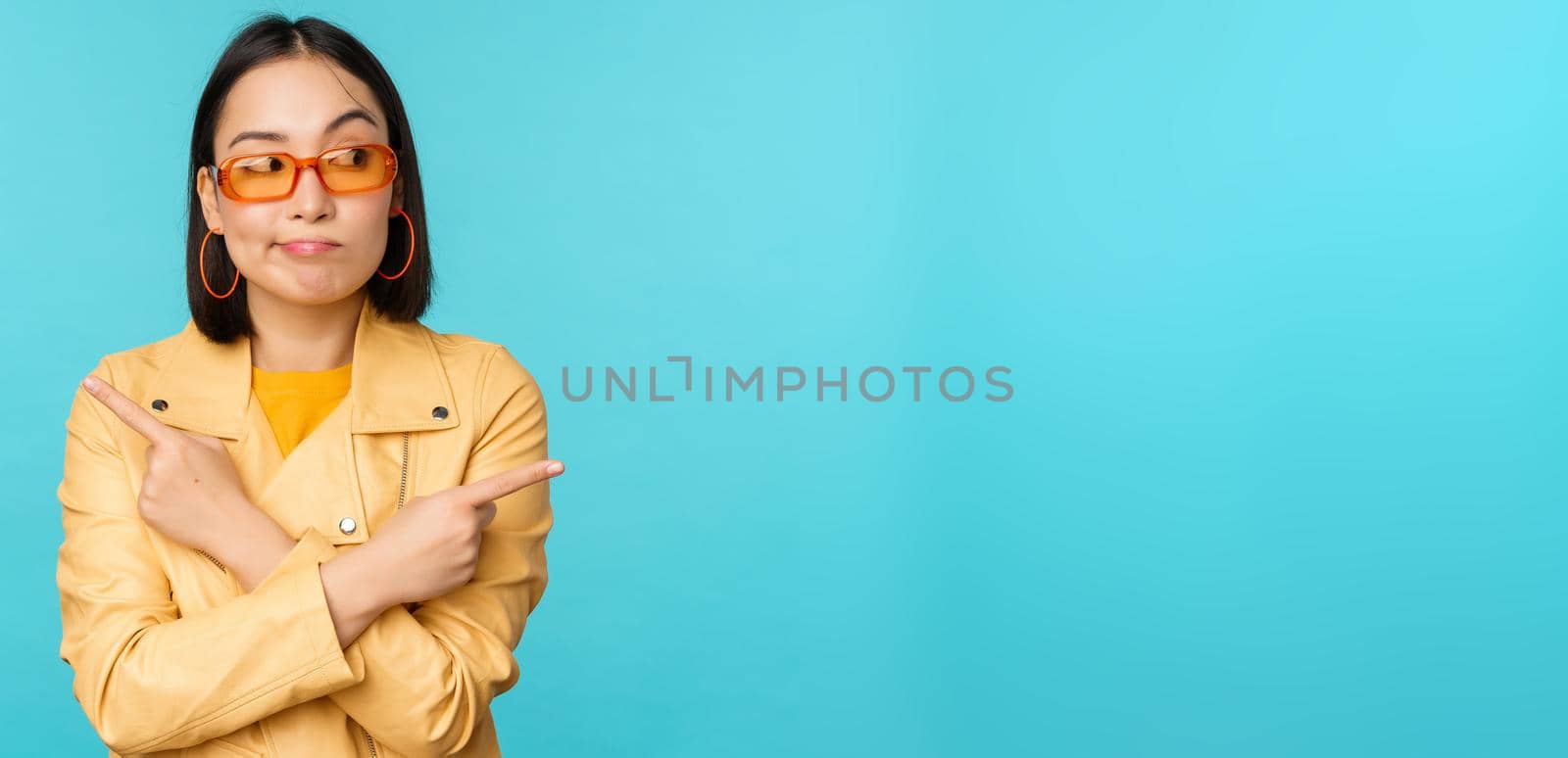 Indecisive asian woman in sunglasses, makes choice between two variants, points sideways, looks puzzled, stands over blue background by Benzoix
