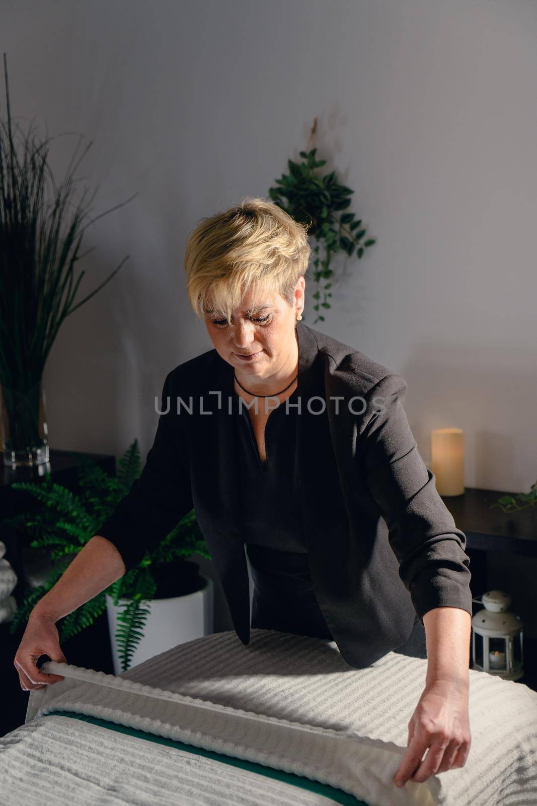 Portrait of a blonde-haired woman, beautician, mature, concentrated and professional, dressed in the black uniform of the company, preparing the massage table to start the day. Relaxing atmosphere and subdued lighting, decoration of plants and candles, background massage table and towels. Vertical