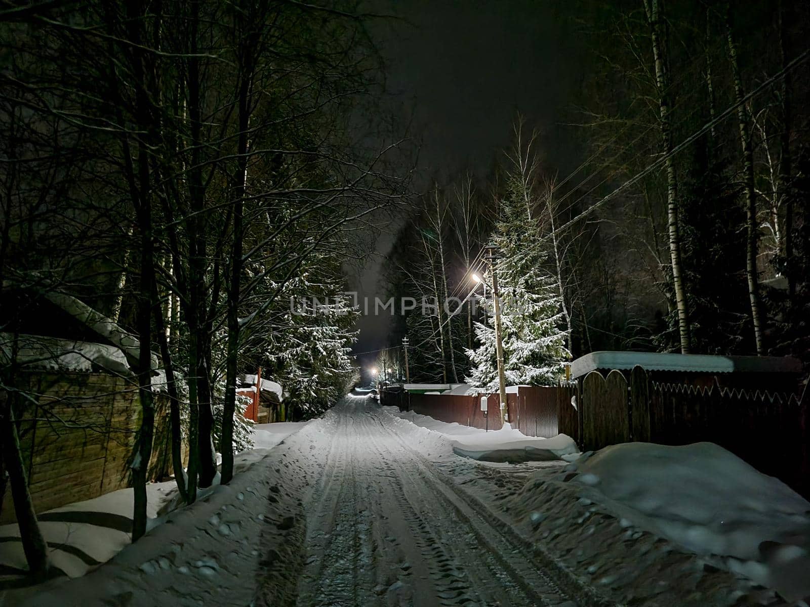 Russian village at night in winter. A road covered with snow and light from the windows of houses by Milanchikov