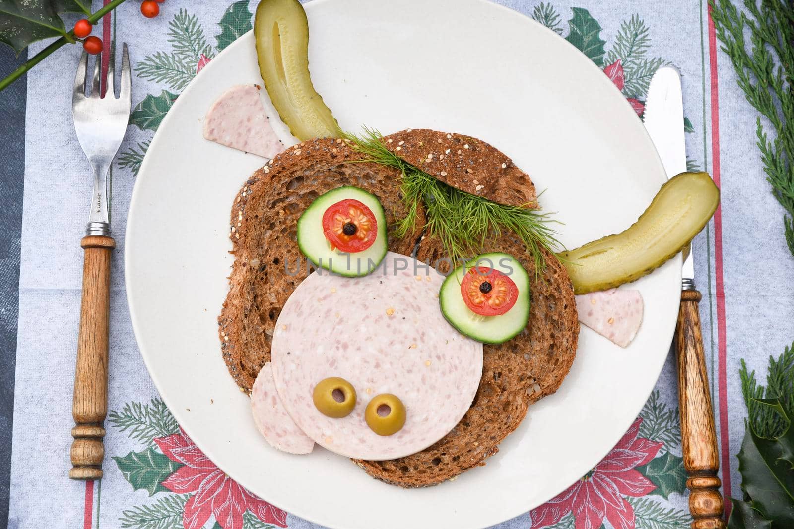 Sandwich in the form of a portrait of a cheerful bull made of dark bread, sausages and vegetables on a plate, on a dark background with a decor of thuja and holly branches