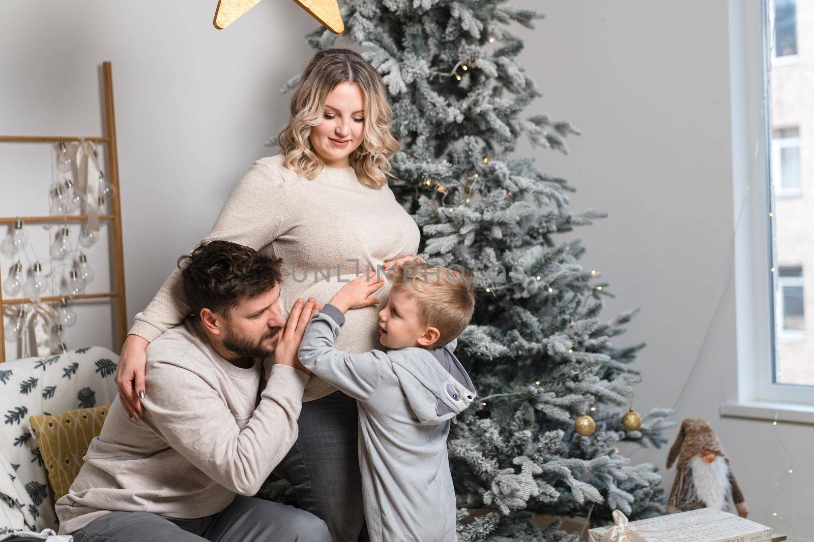 Christmas Family Happiness Portrait of dad, pregnant mom and little son sitting armchair at home near Christmas tree hug smile European young adult family holiday morning