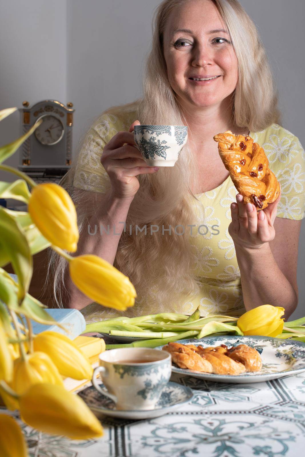 Drinking coffee and tea, a middle-aged blonde woman with long hair drinks tea from a vintage cup and eats puff pastry with pecans on the background of yellow tulips and books on the table