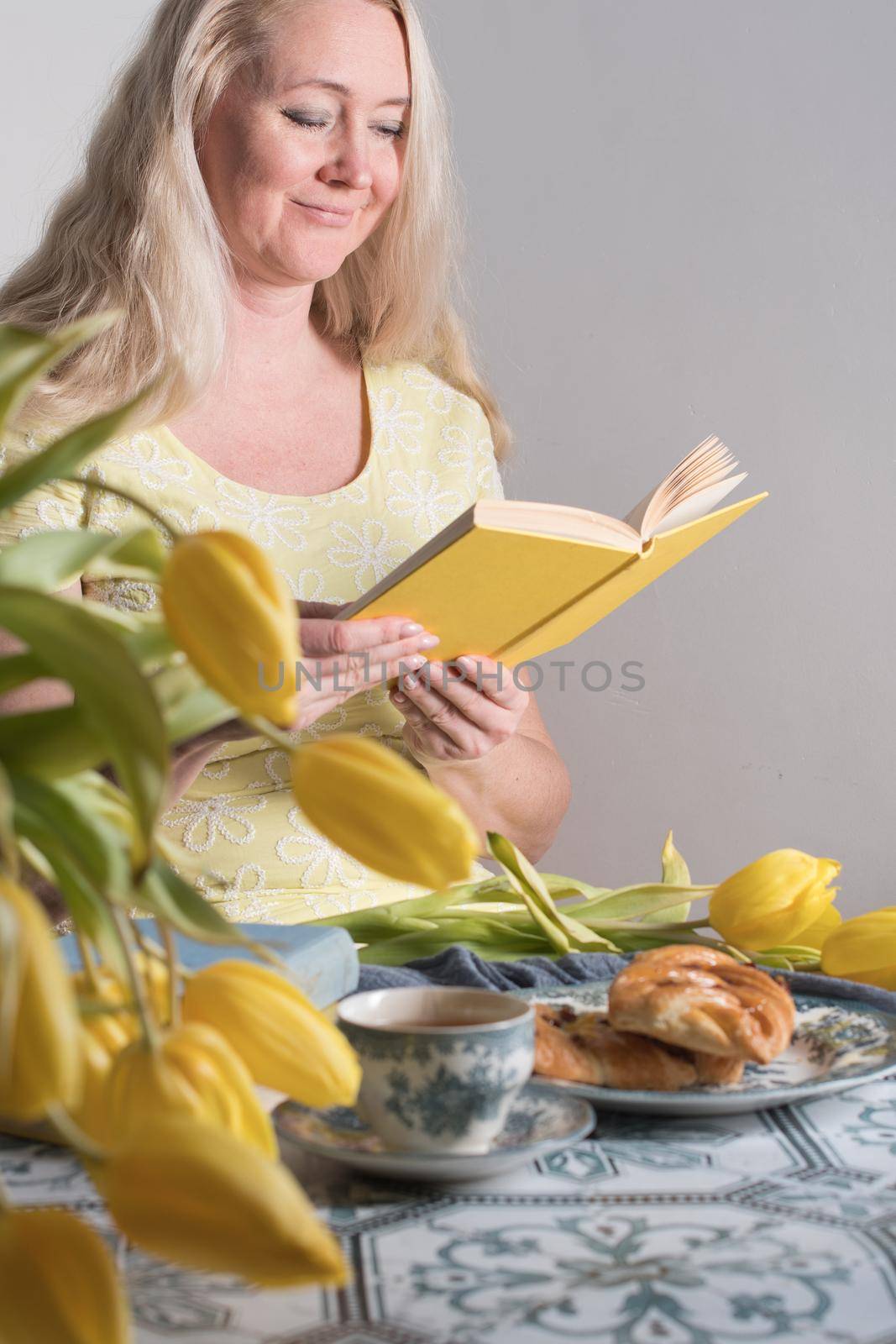 Drinking coffee and tea, a middle-aged blonde woman with long hair drinks tea from a vintage cup and eats puff pastry with pecans on the background of yellow tulips and books on the table