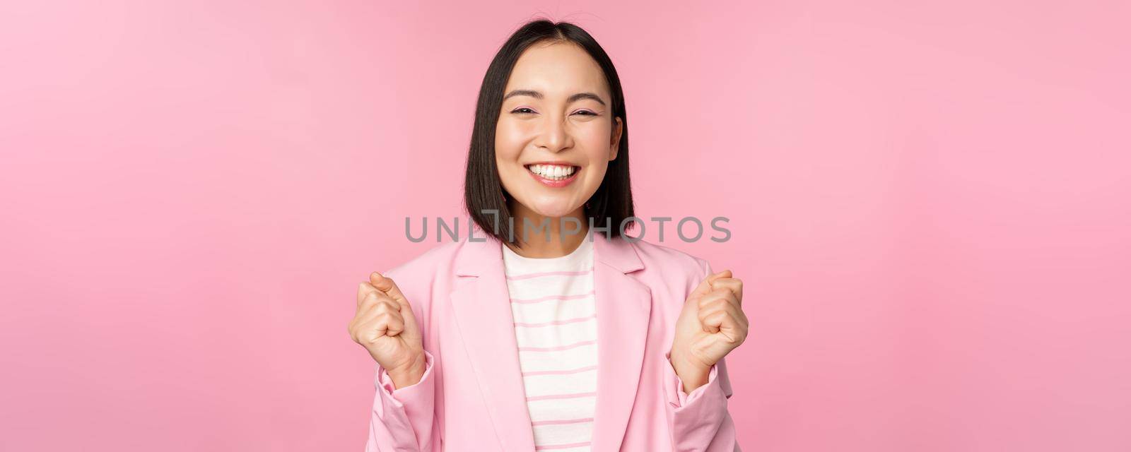 Enthusiastic saleswoman, asian corporate woman say yes, achieve goal and celebrating, triumphing, looking with rejoice and smiling, standing over pink background by Benzoix