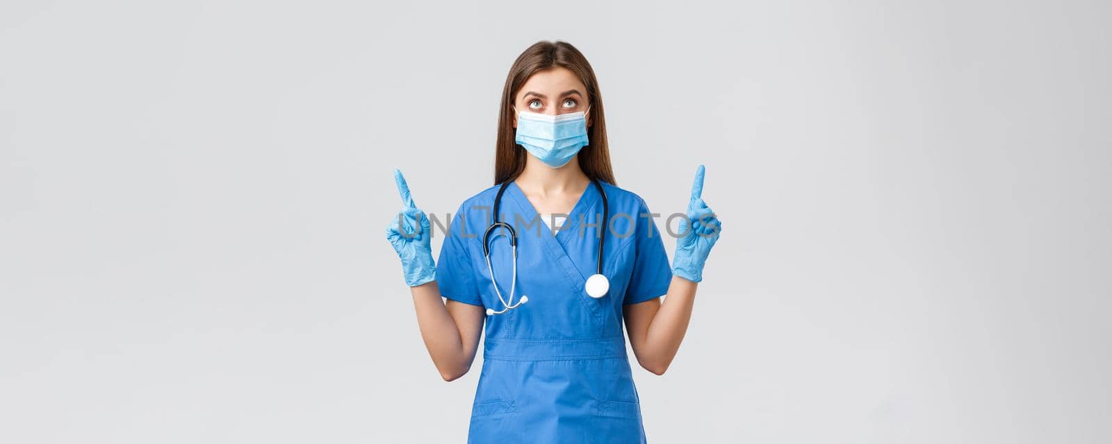 Covid-19, preventing virus, health, healthcare workers and quarantine concept. Curious female doctor or nurse in blue scrubs, medical mask reading banner, pointing fingers up.