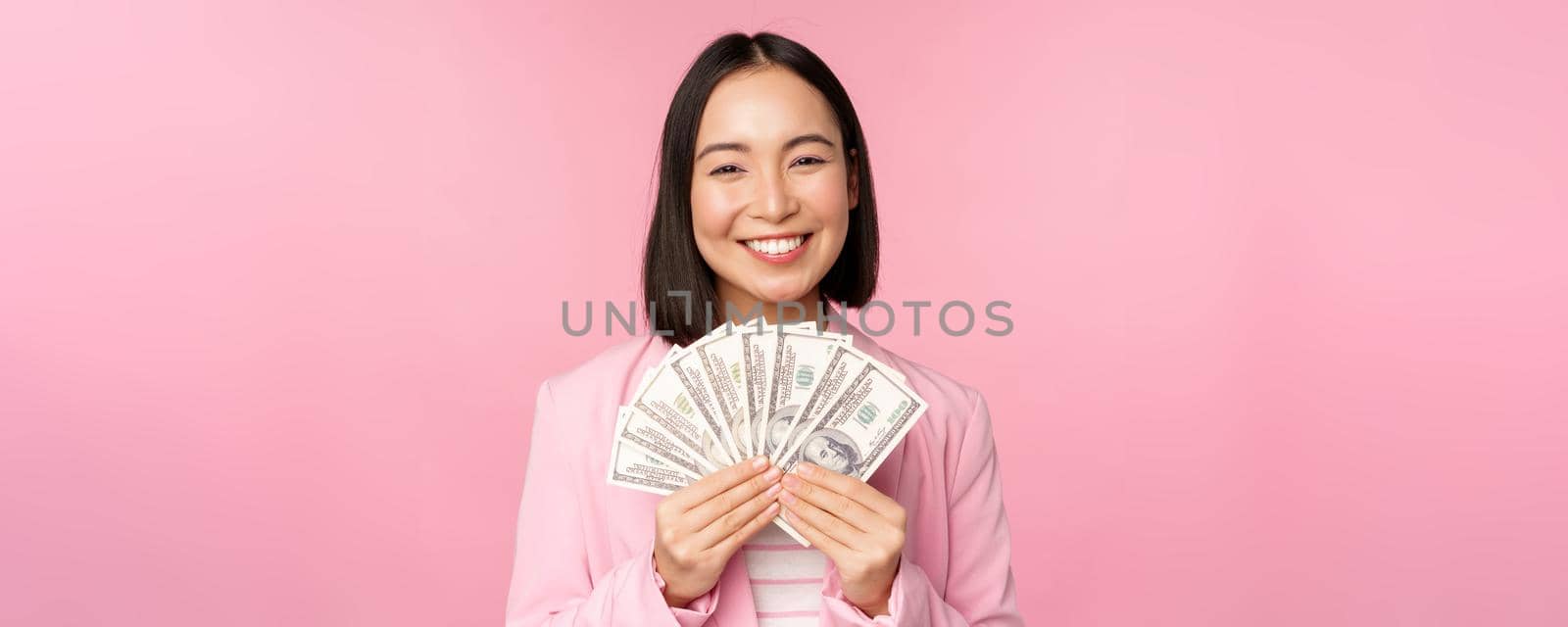 Finance, microcredit and people concept. Happy smiling asian businesswoman showing dollars money, standing in suit against pink background.