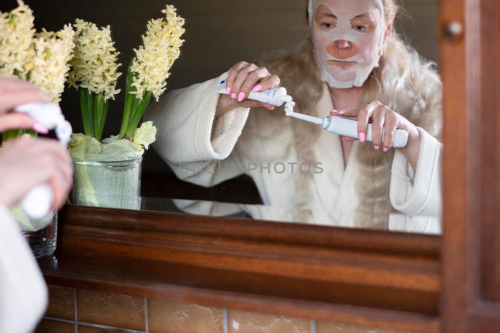 a middle-aged woman in a refreshing mask is busy with morning hygiene, she applies paste on an electric toothbrush, High quality photo