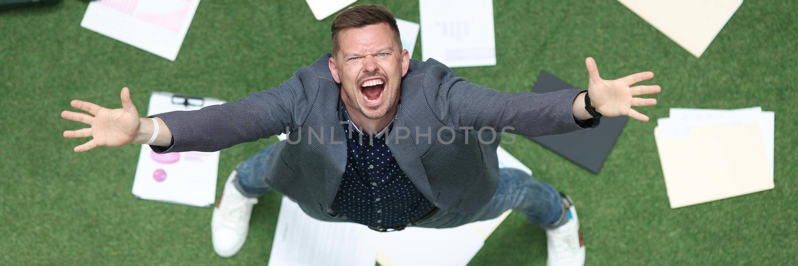 Man in suit throwing papers on green grass and yelling with his hands up top view by kuprevich